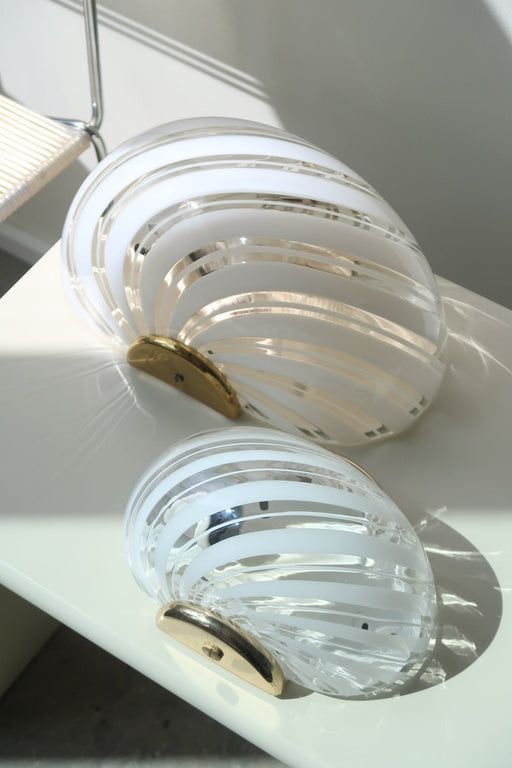 Sæt á 2 stk vintage Murano væglamper i glas med swirl mønster og beslag med messingoverflade. Perfekt størrelse til din entré, i køkkenet, på badeværelset eller som læselampe i soveværelset. Super nemme at montere. Mundblæst i Italien, 1960/70erne og varierer i udtrykket med alderssvarende patina.  Størrelse 22x15 cm. 